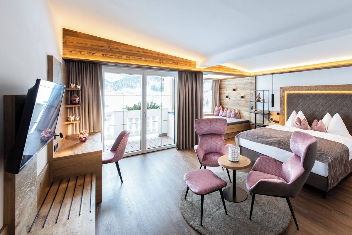 A modern hotel room with a large bed, a TV, a seating area with chairs, and a view of the outdoors.