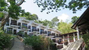A multi-story hotel with a stone staircase leading to the entrance, surrounded by lush greenery and a clear sky. - Aiyanar Beach and Dive Resort (Mabini)