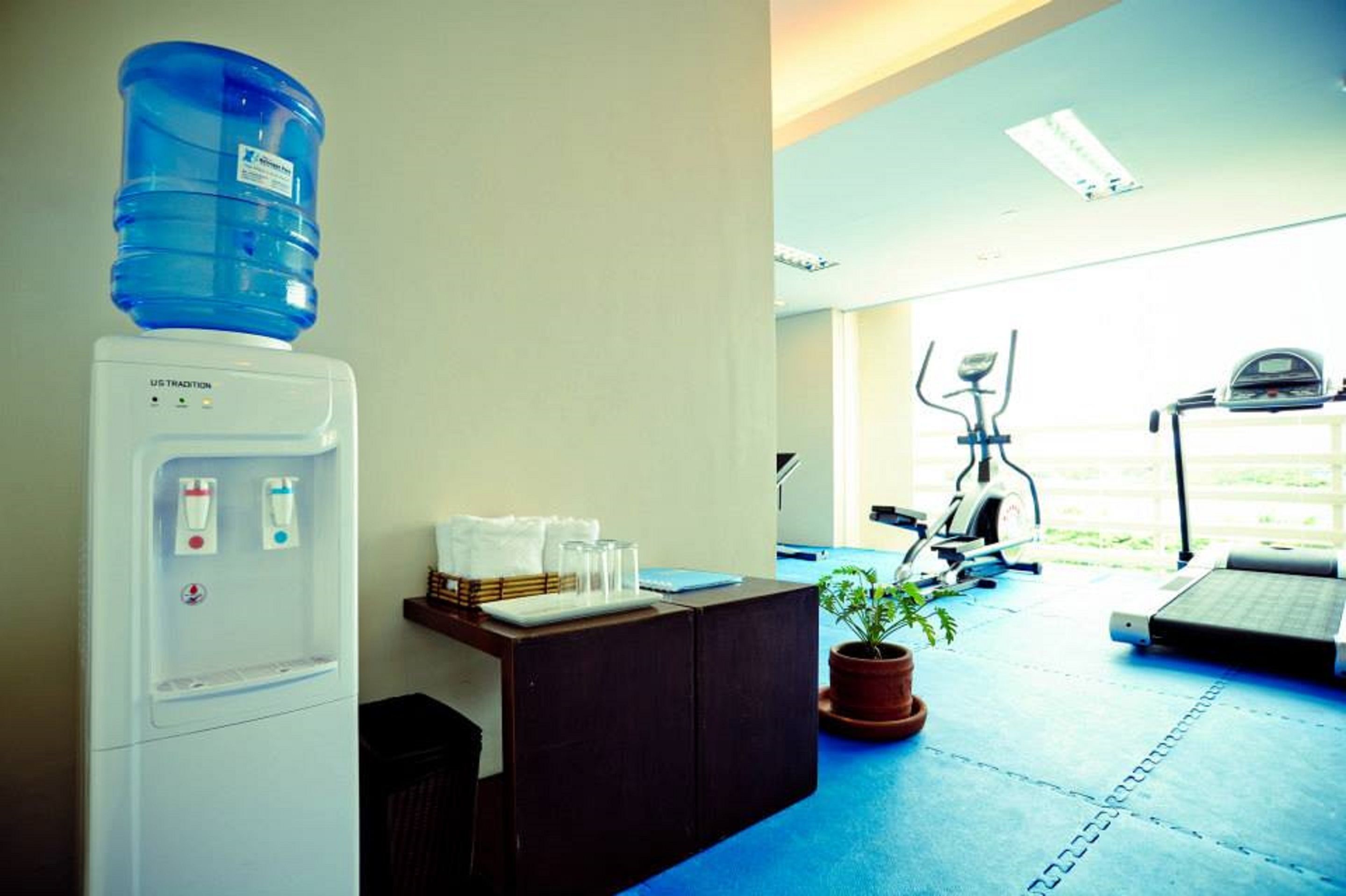 A reception area with a water dispenser, a desk with a plant, and exercise equipment in the background.
