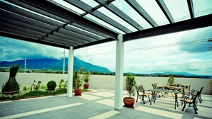 Une terrasse extérieure couverte, avec une table et des chaises, des plantes en pot et une vue sur les montagnes.