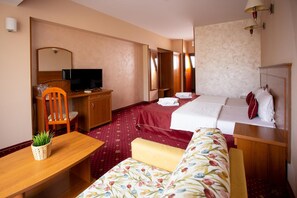 A hotel room with a bed, a desk with a television, a chair, a small table with a plant, and a sofa.