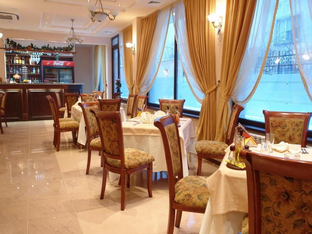 A restaurant with multiple tables and chairs, a bar area with a Coca-Cola refrigerator, and large windows with curtains.
