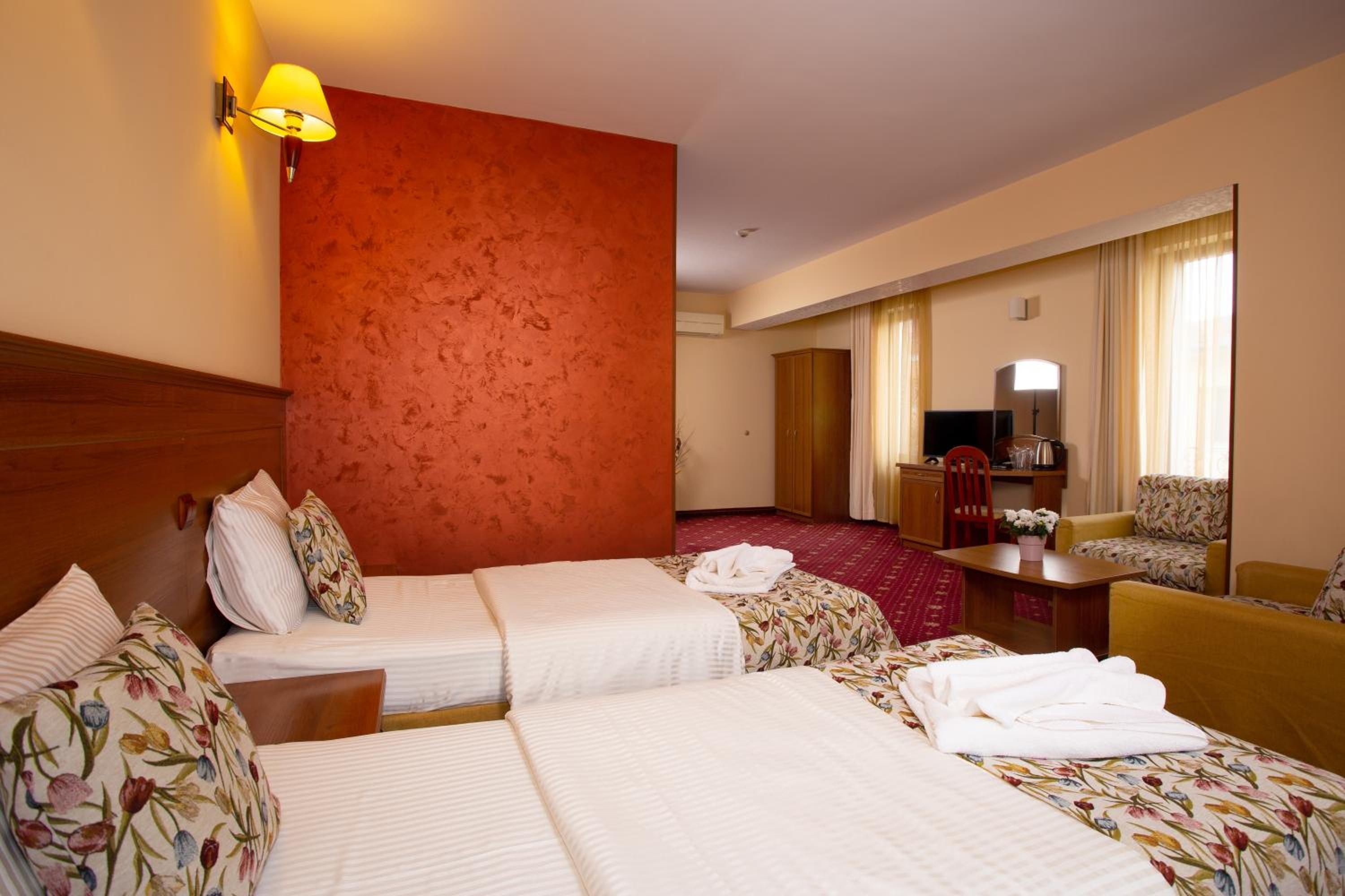 A hotel room with two beds, a sitting area with a sofa and armchair, a desk, and a large window with curtains.