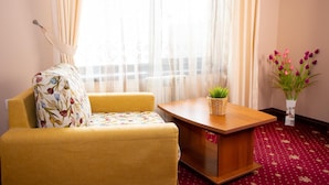 Een kamer met een gele fauteuil, een klein houten tafeltje met een rieten mand, en een vaas met bloemen.