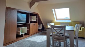 Een kamer met een televisie, houten kasten, een gele bank en een eettafel met stoelen.