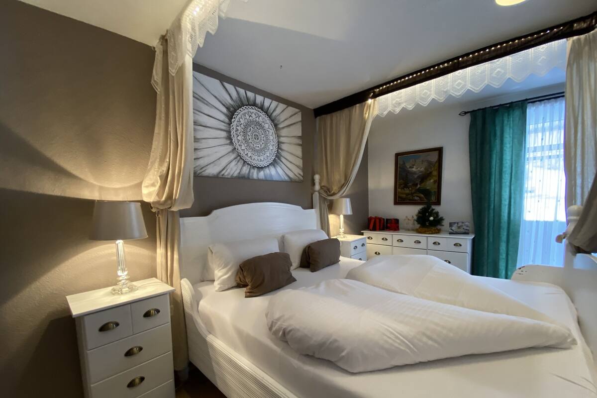 Un dormitorio con una cama grande, una mesita de noche, un aparador, un adorno en la pared y una ventana con cortinas.