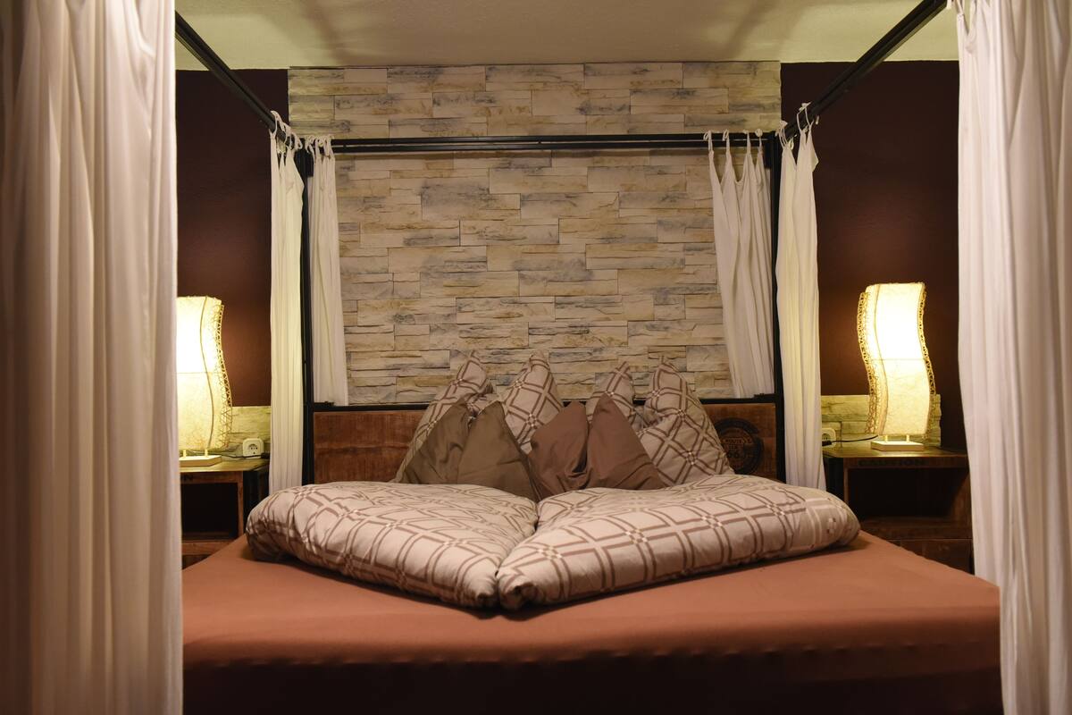 Una cama con cabecera, dos mesitas de noche con lámparas y una pared con diseño de piedra al fondo.