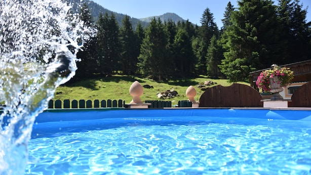 A swimming pool with clear blue water, surrounded by a wooden fence and a backdrop of green hills and trees. - Apartment Fernerkogel (Gries im Sellrain)