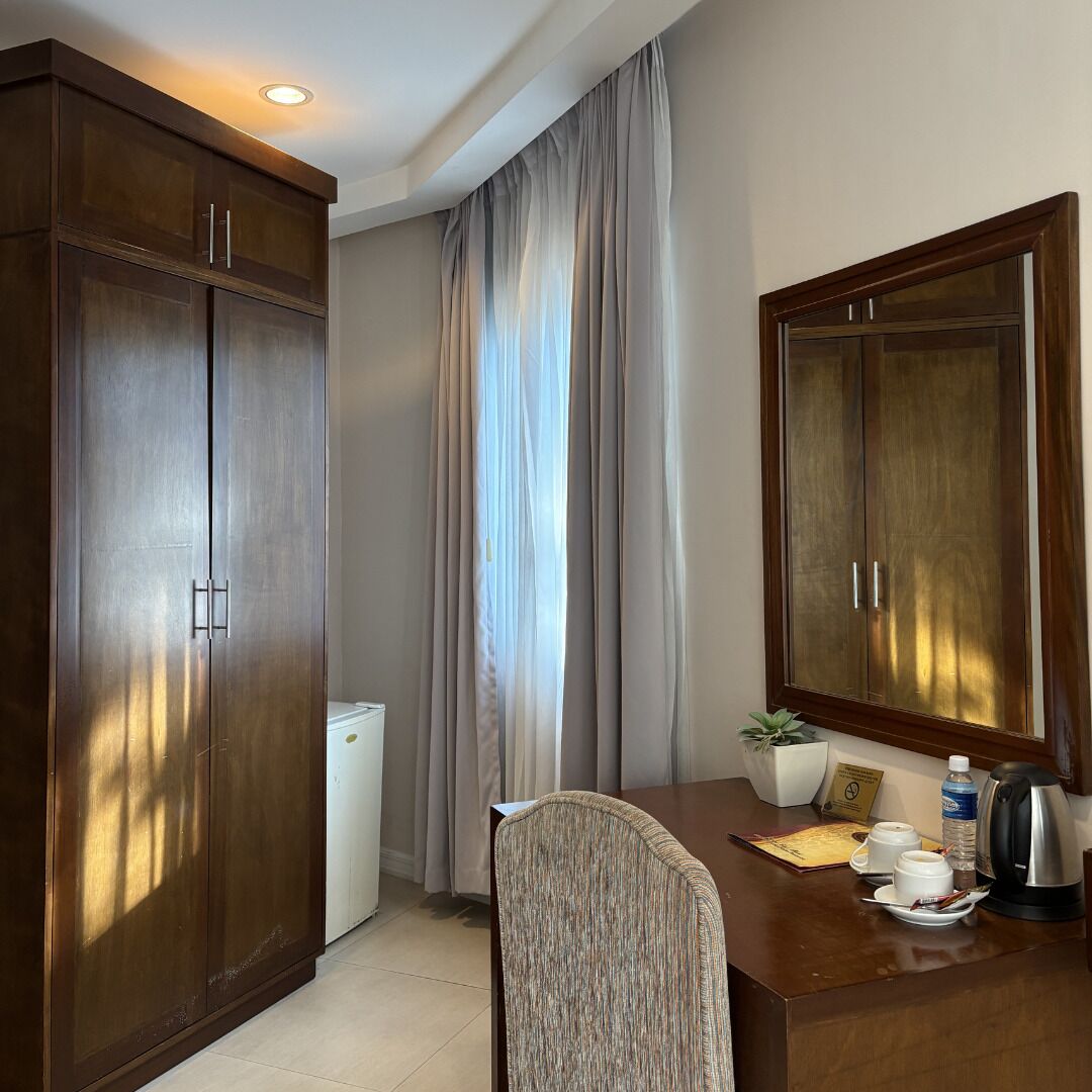 A hotel room with a wooden desk, a chair, a small refrigerator, a potted plant, a coffee maker, and a large mirror.