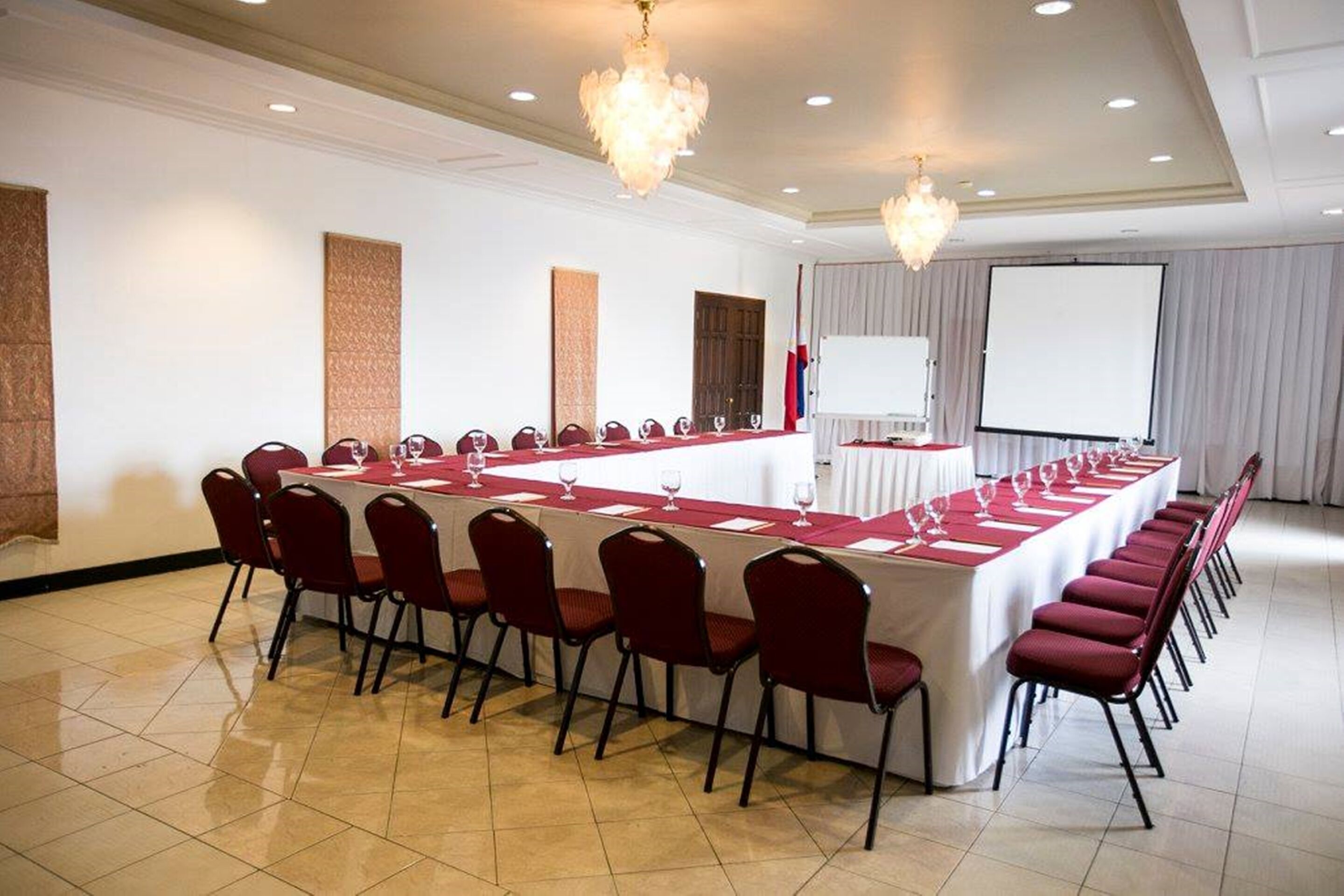 Un salón de conferencias con una larga mesa, sillas rojas, una pizarra y una pantalla de proyector.