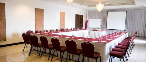 Un salón de conferencias con una larga mesa, sillas rojas, una pizarra y una pantalla de proyector.