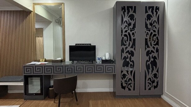 A hotel room with a bed, a desk with a TV, a chair, and a wardrobe. - Queen Margarette Hotel - Main (Lucena)