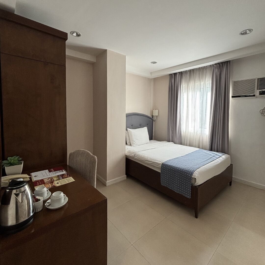 A hotel room with a bed, a desk with a kettle and cups, a chair, and a window with curtains.