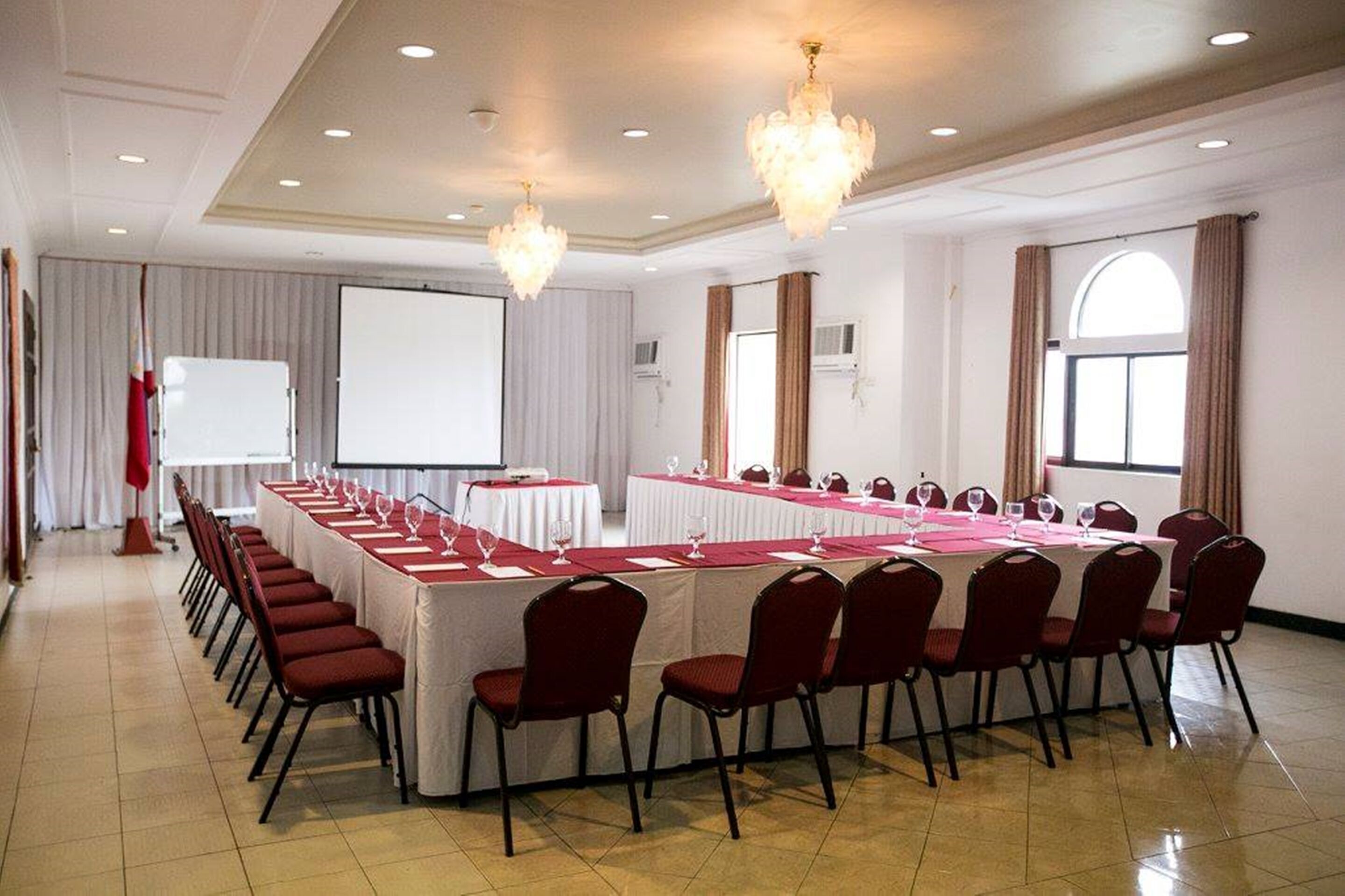 Un salón de conferencias con una larga mesa, sillas rojas, candelabros y una pantalla para proyector.