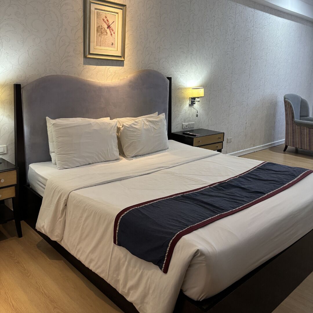 A neatly made bed with white linens and a dark blue accent, a bedside table with a lamp, and a chair in a room with patterned wallpaper.