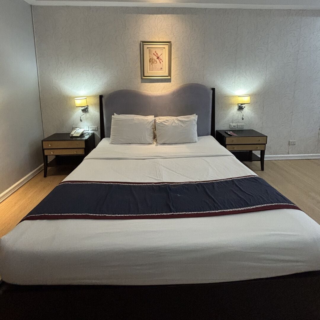 A neatly made bed with white linens and a dark blanket, flanked by two bedside tables with lamps, in a room with patterned wallpaper and a framed picture on the wall.