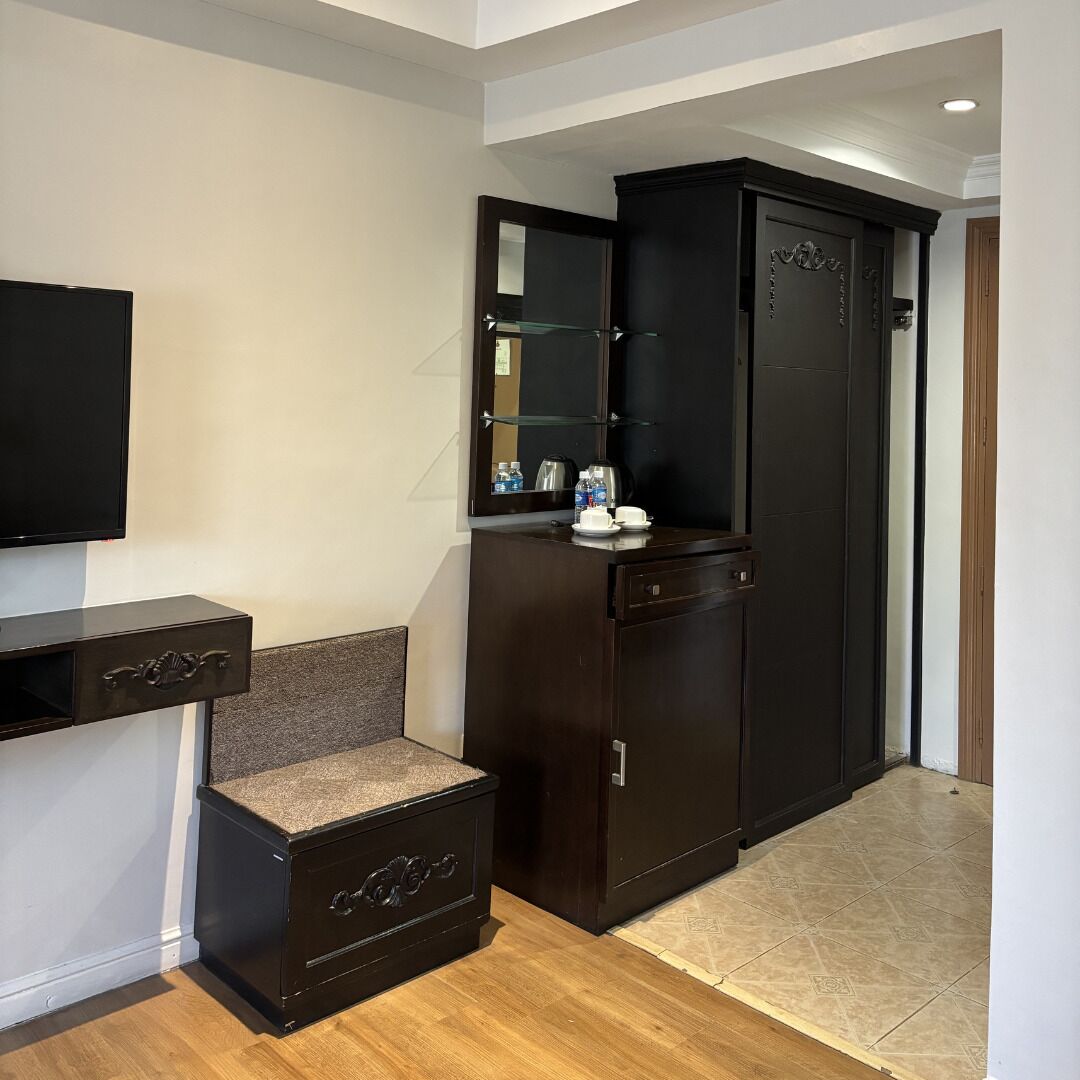 A hotel room with a dark wooden wardrobe, a small seating area with a cushioned bench, a flat-screen TV mounted on the wall, and a tiled floor.