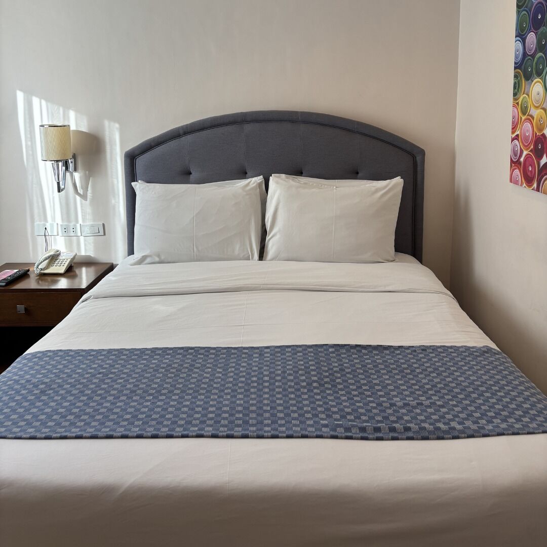 A neatly made bed with a patterned bedspread, a bedside telephone, and a wall-mounted lamp.