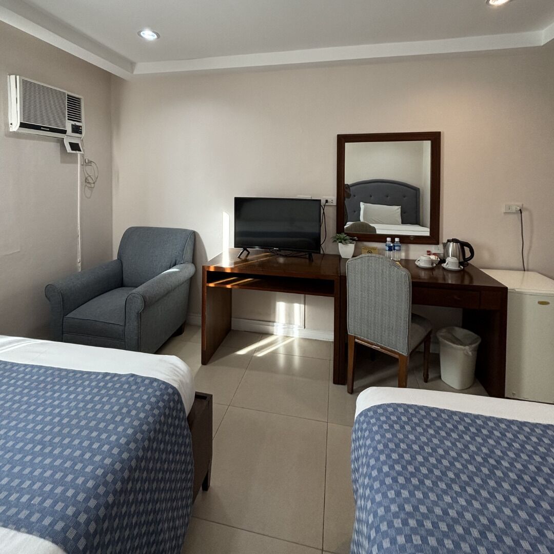 A hotel room with two beds, a desk with a television, a chair, a small refrigerator, and a trash bin.