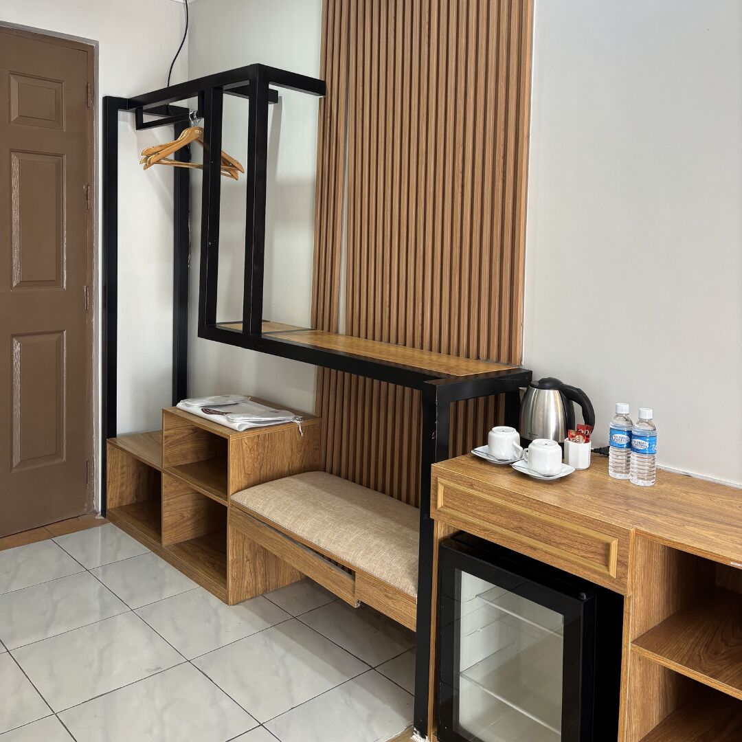 A compact room with a wooden bench, a small table with a kettle, cups, and water bottles, and a wall-mounted shelf with a hanging garment.