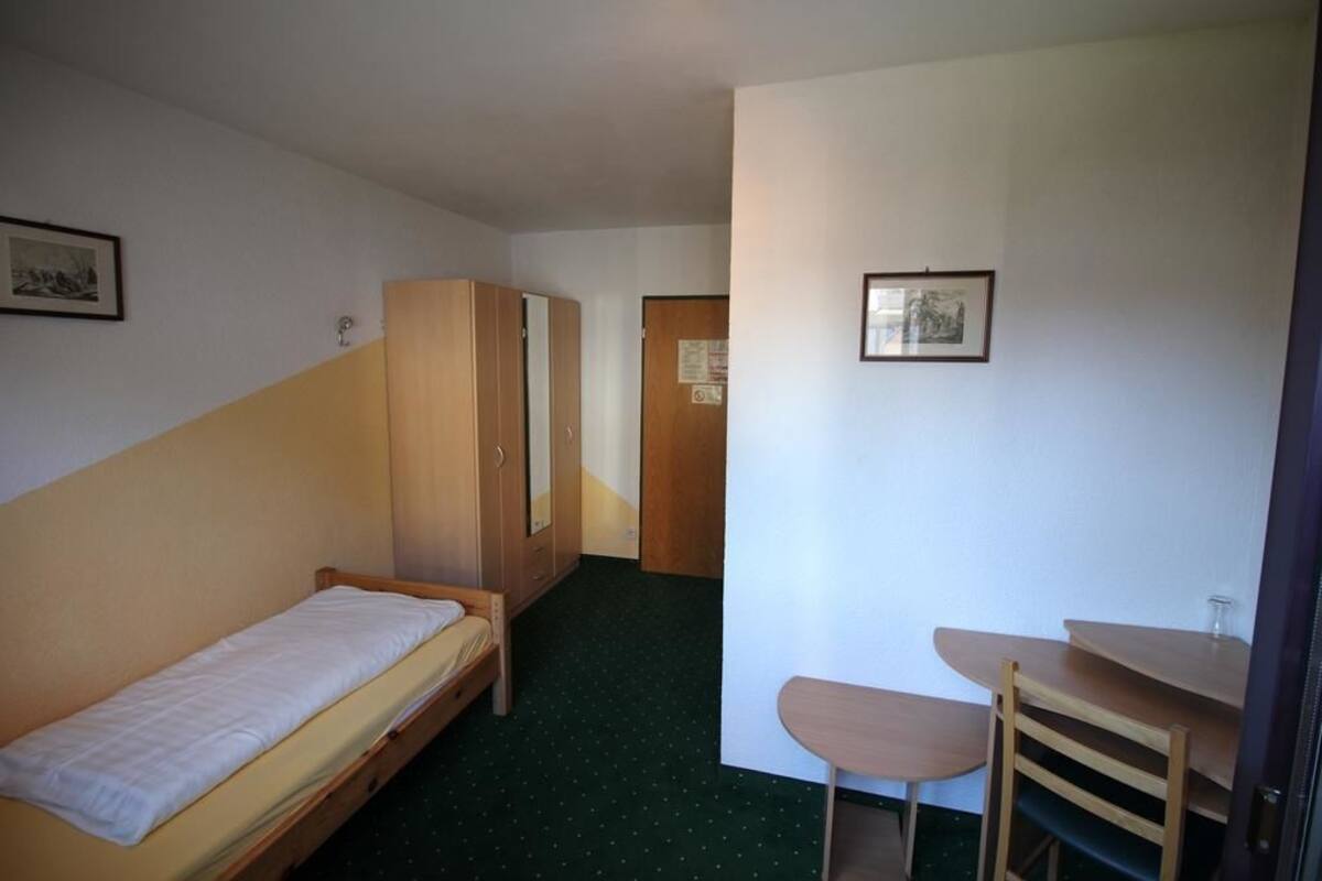Een kleine, compacte kamer met een enkel bed, een houten garderobe, een tafeltje met een stoel en een deur naar een andere kamer.