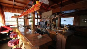 Uma área de bar com balcão de madeira, banquetas vermelhas e diversas garrafas e copos.