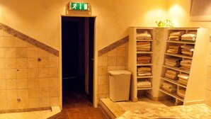 Um corredor com piso ladrilhado, uma porta com placa de saída de emergência, uma cesta de vime e uma prateleira com toalhas dobradas.