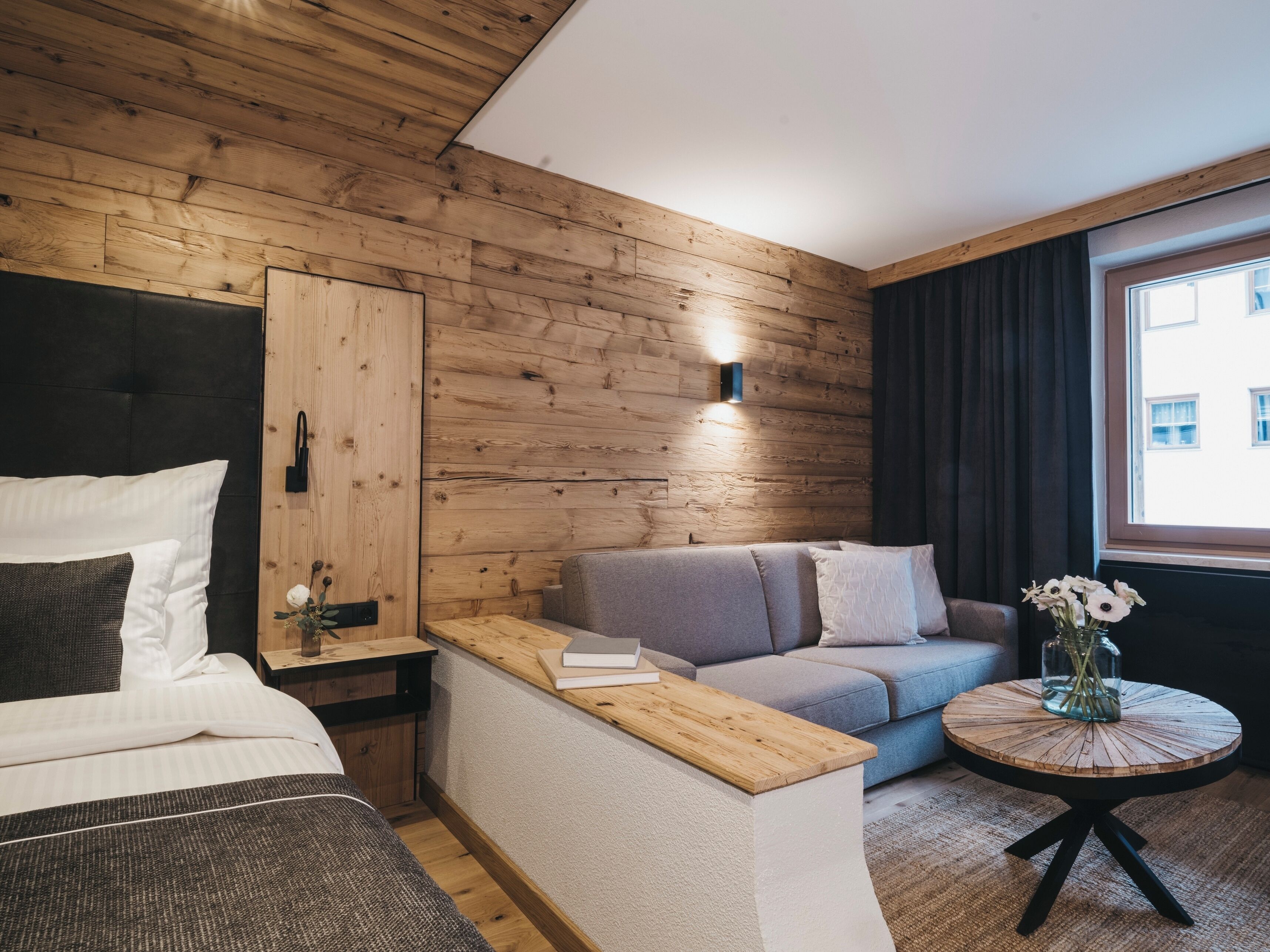 Una habitació d'hotel moderna amb una paret de fusta, un llit, un sofà, una taula petita i una taula rodona de fusta amb un ram de flors.