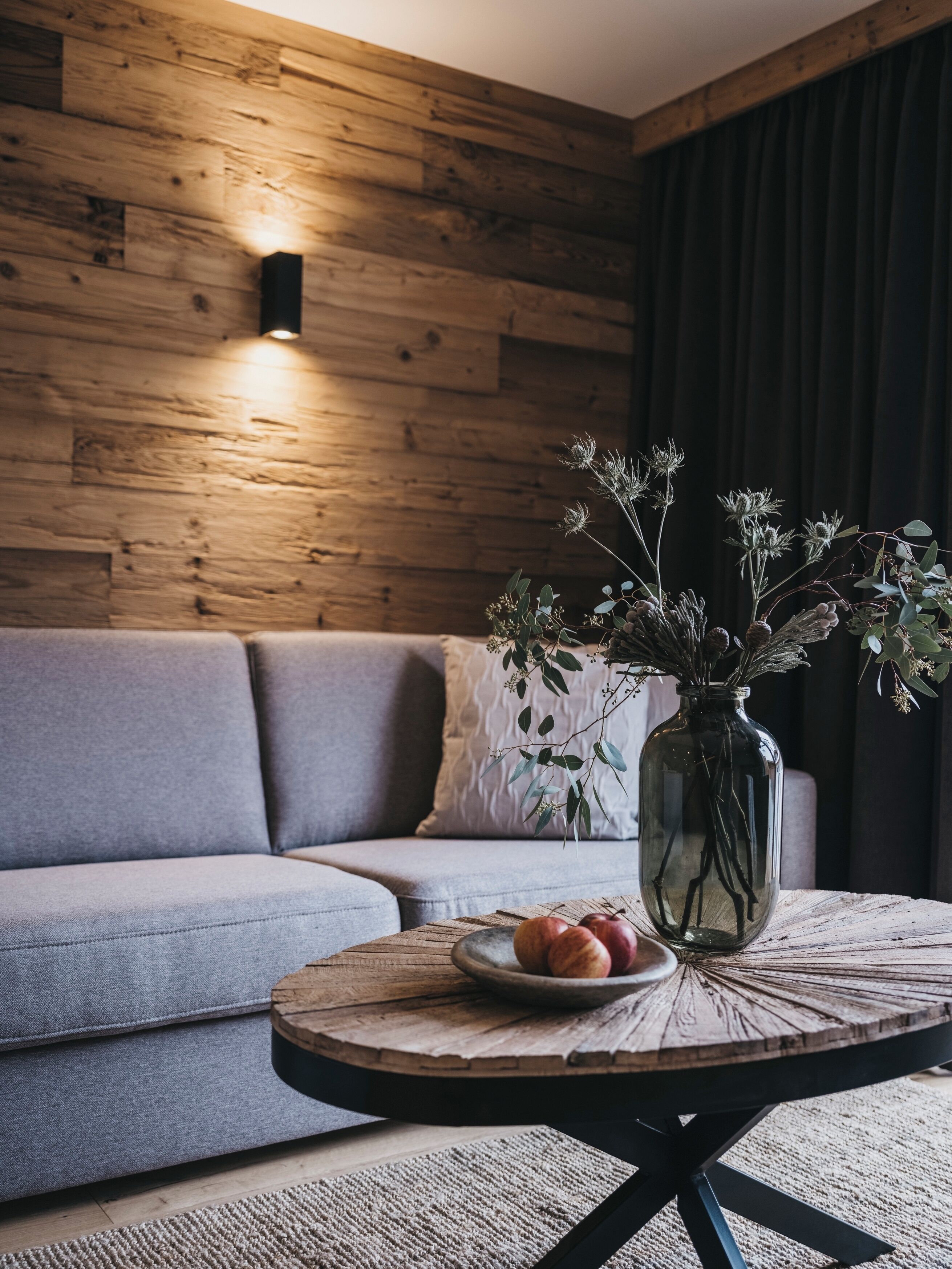 A modern living room with a wooden wall, grey sofa, and a round coffee table with a vase of dried flowers and a plate of fruit.