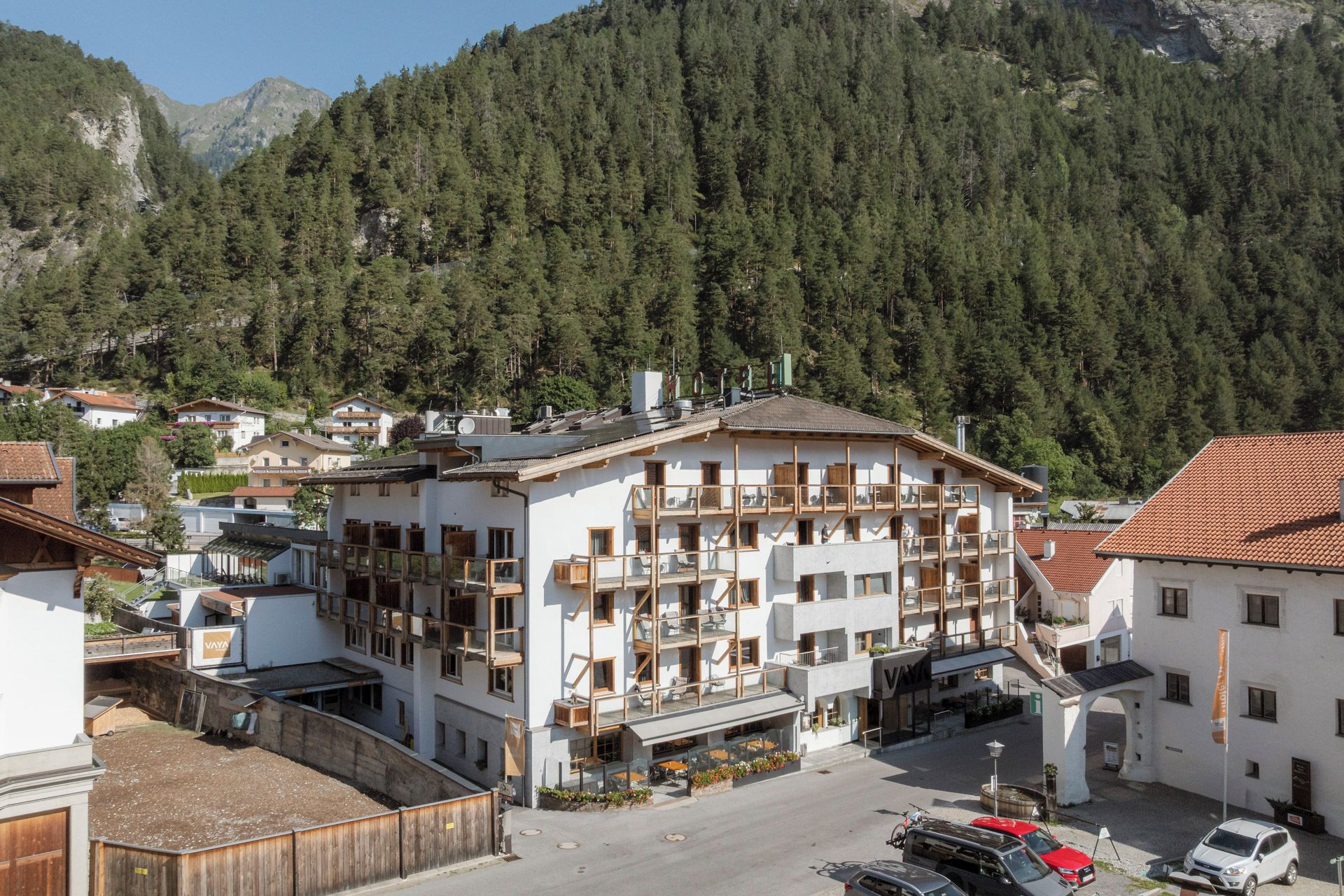 A multi-story building with balconies, a parking area, and a mountainous backdrop.