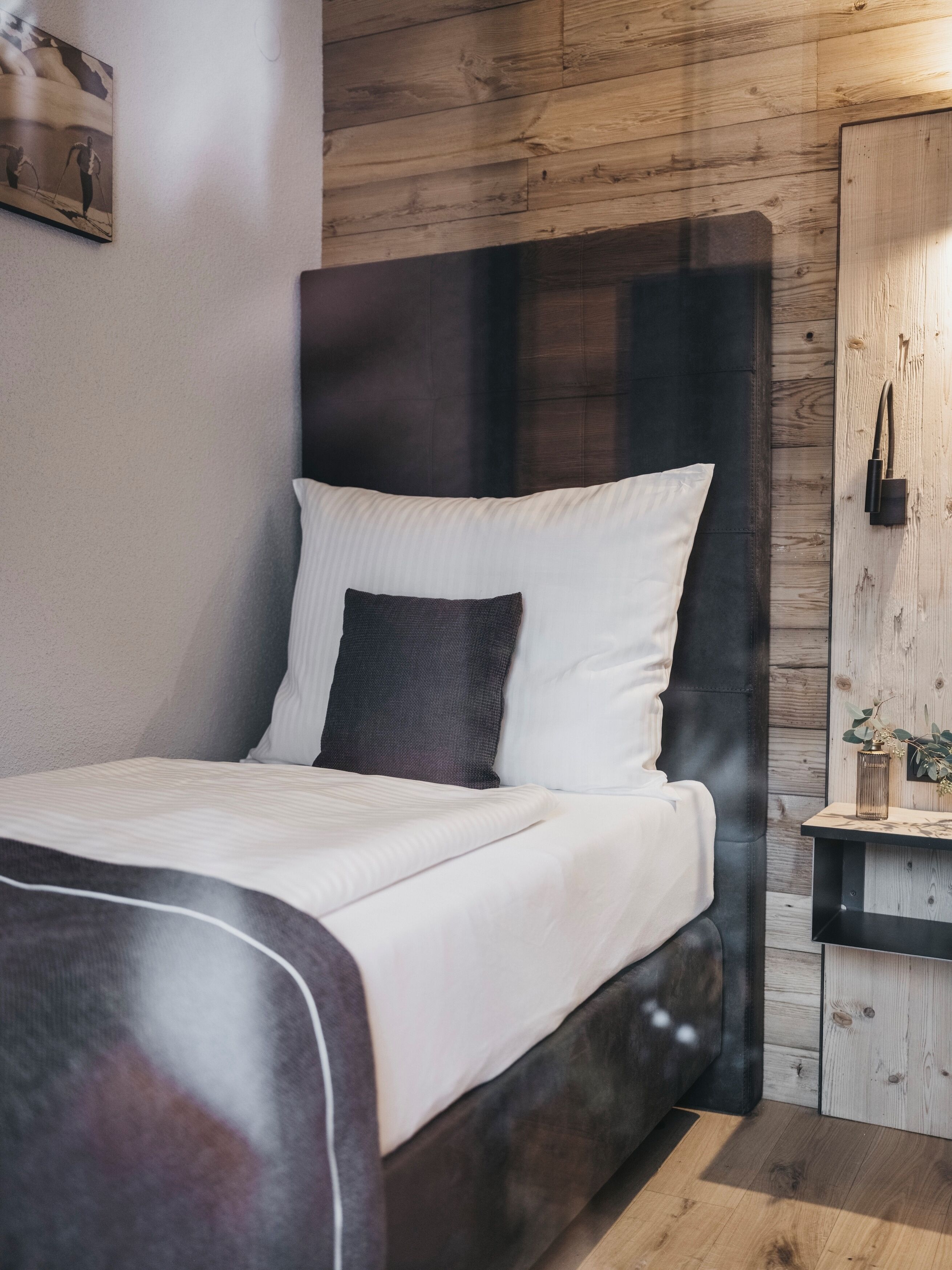 A bed with white and dark pillows, a wooden headboard, and a small bedside table with a plant.