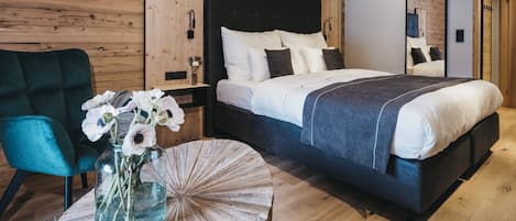 A modern bedroom with a wooden ceiling, a large bed with white and dark bedding, a teal armchair, and a vase with flowers on a round wooden table.