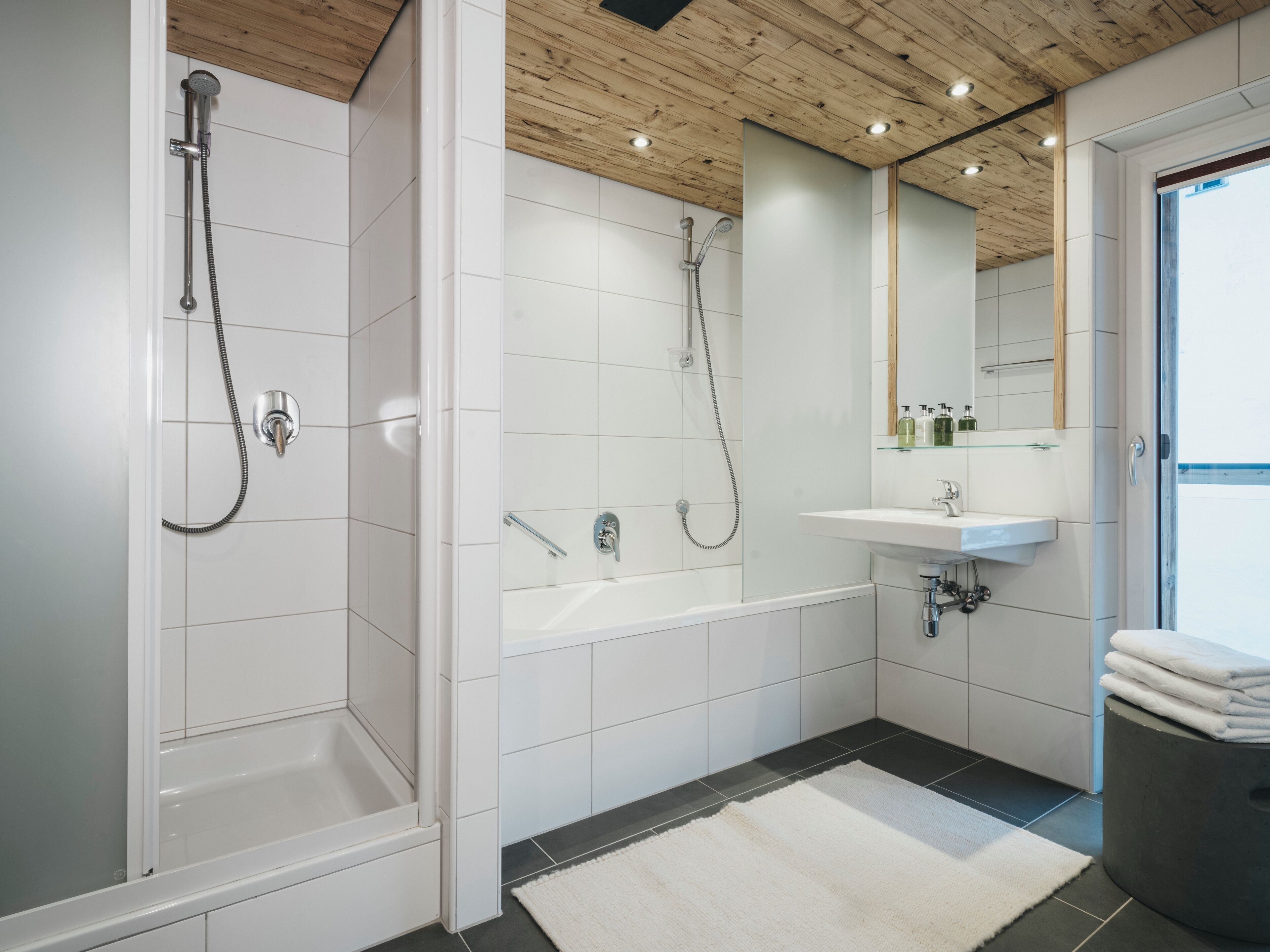 A modern bathroom with white tiles, a wooden ceiling, a bathtub, a shower, and a sink with a mirror above it.