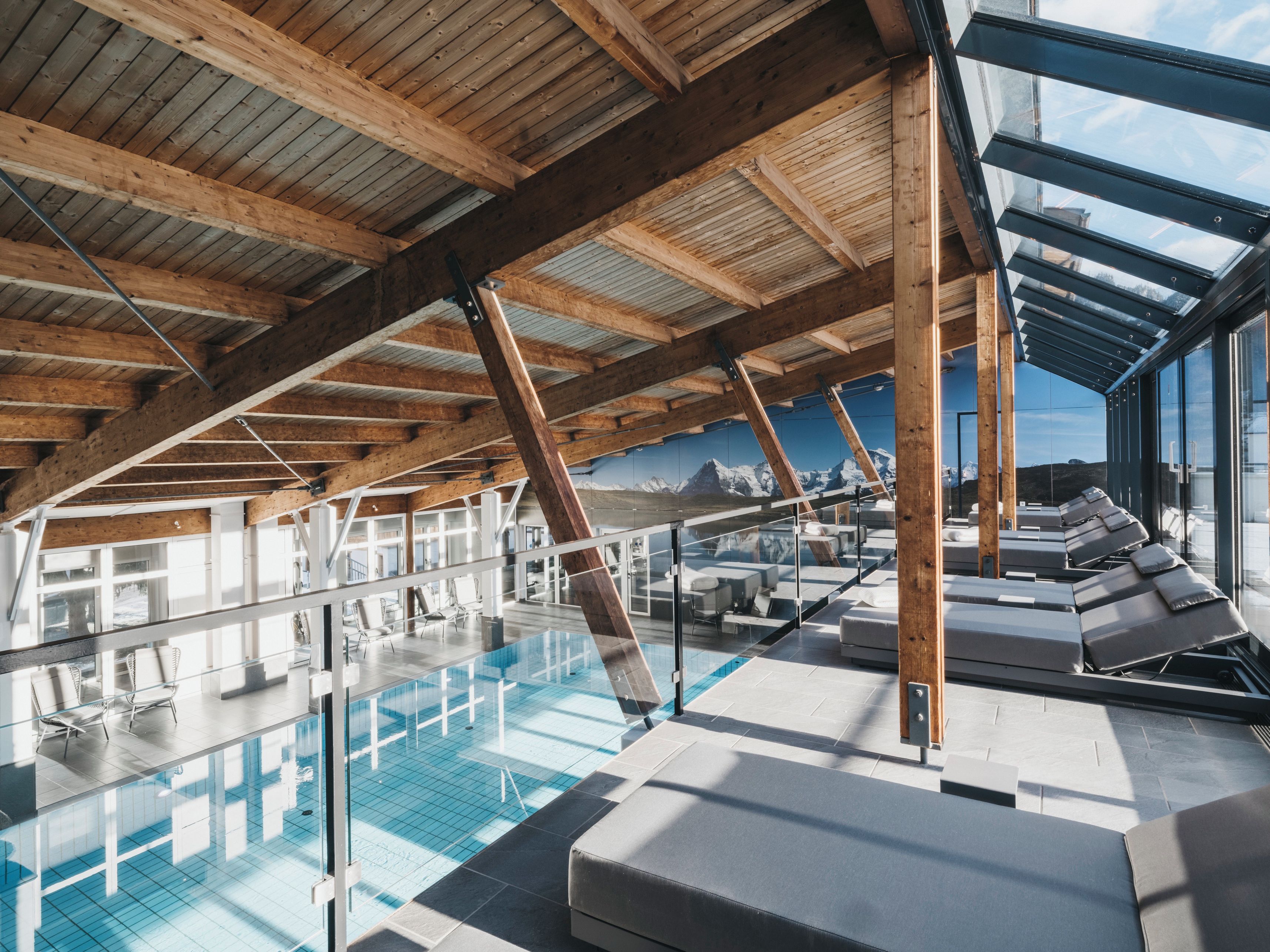 Un espace piscine intérieur moderne doté d’un plafond en verre, de poutres en bois et de coins salon confortables.