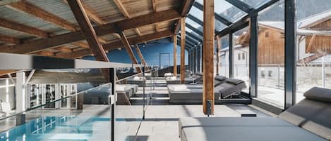 A modern indoor pool area with clear glass walls, wooden beams, and lounge seating.