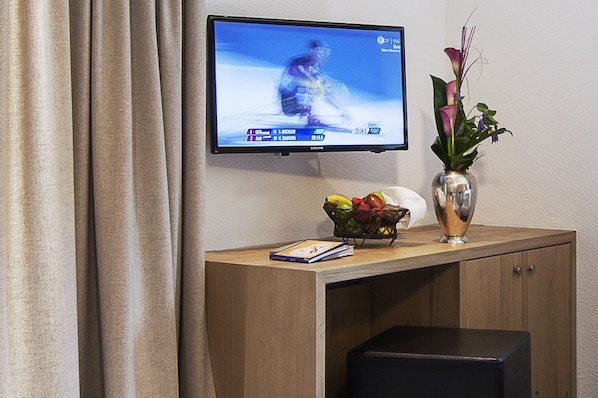 A television mounted on a wall displaying a sports event, a vase with flowers, a fruit bowl, and a book on a wooden desk. - Das Lafairs (Pfunds)