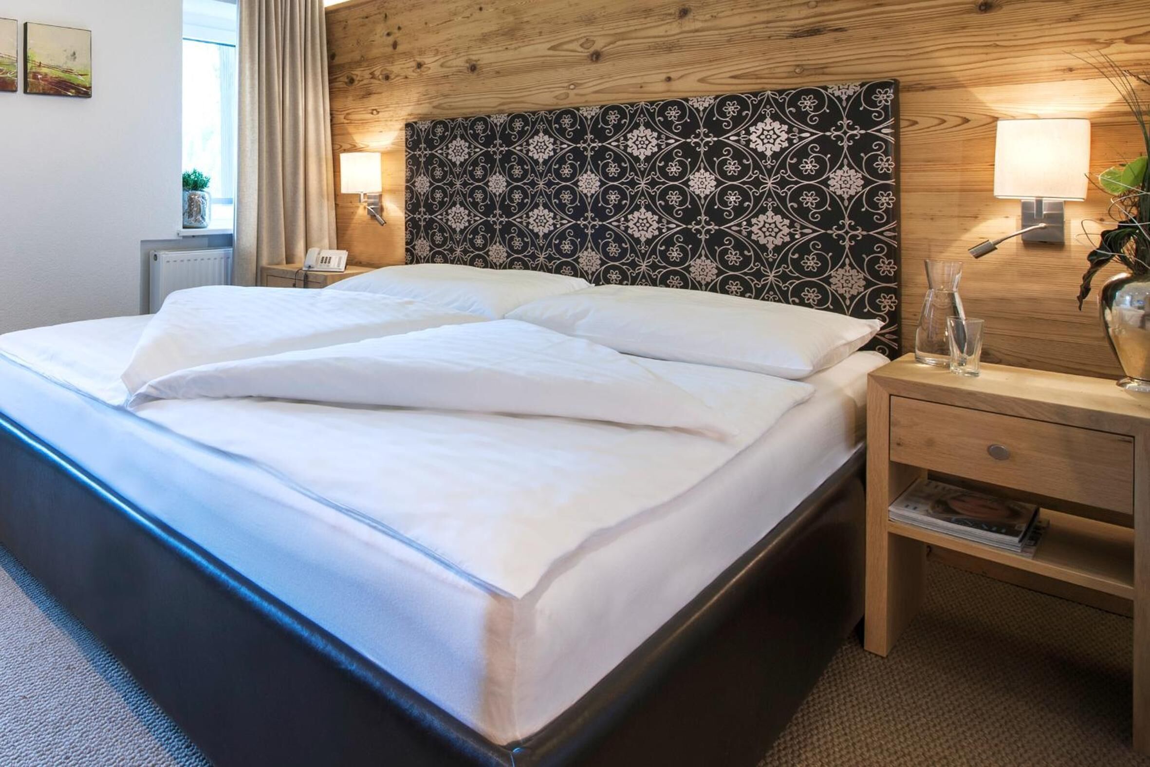 A neatly made bed with white linens in a room with wooden paneling and a patterned headboard. There is a bedside table with a lamp and a small plant.