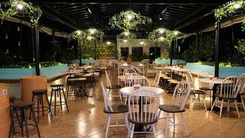 An outdoor restaurant with multiple tables and chairs, surrounded by greenery and a tiled floor.