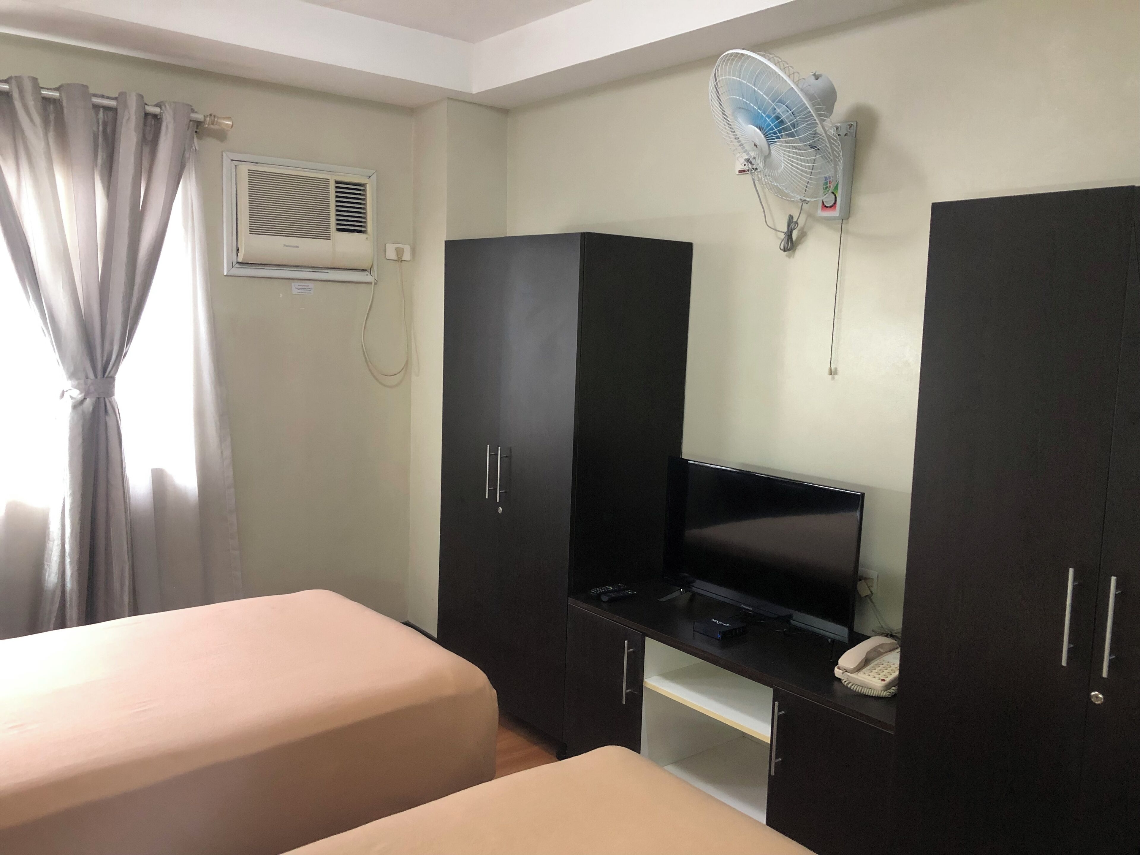 A hotel room with two beds, a TV, a desk, a phone, a fan, and a window with curtains.