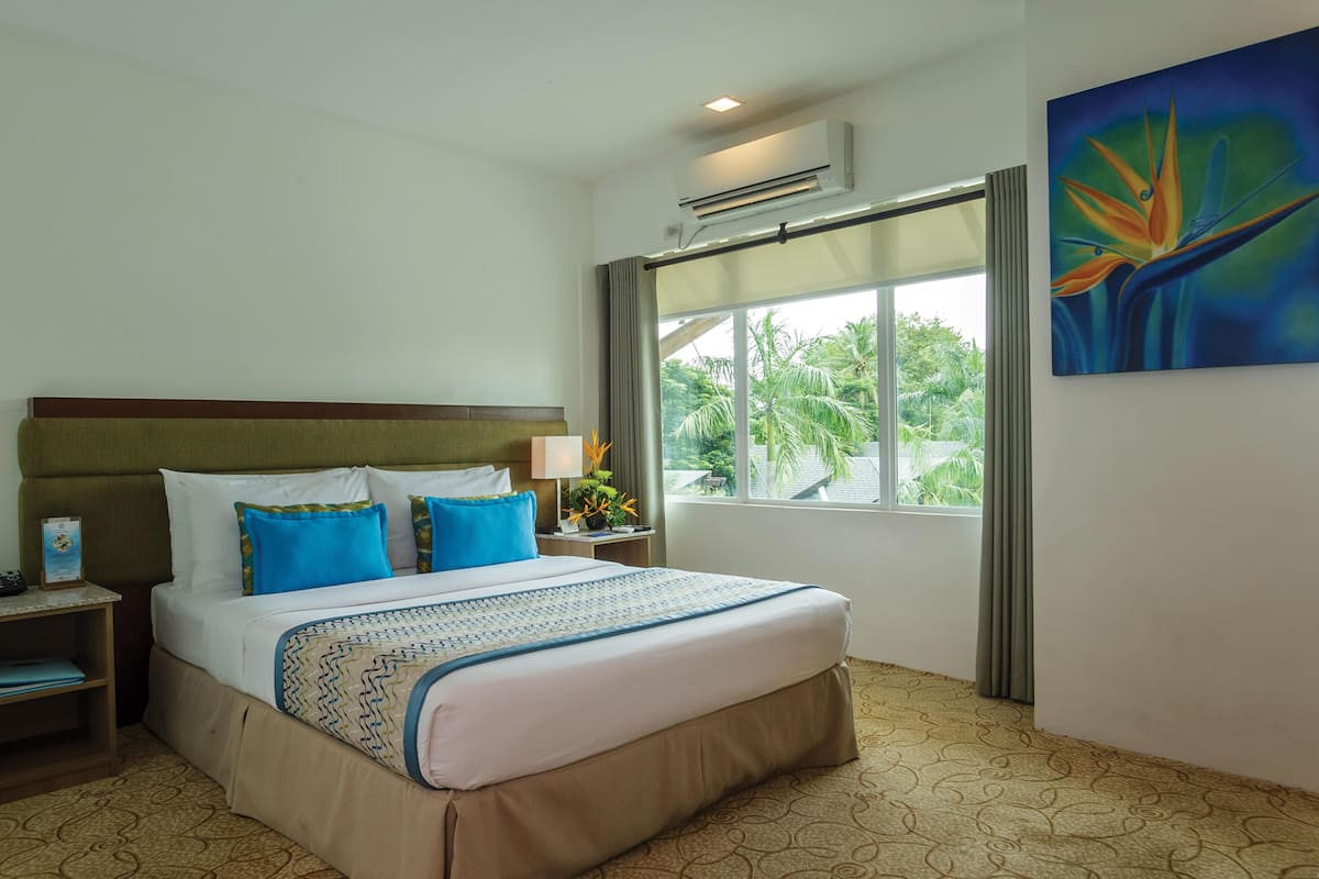 A hotel room with a large bed, a nightstand, a lamp, an air conditioning unit, a window with a view of palm trees, and a painting on the wall.