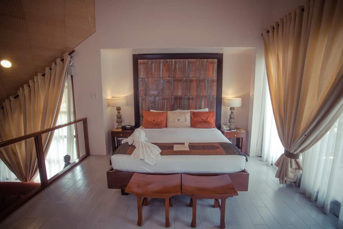 A hotel room with a large bed, wooden headboard, two bedside tables with lamps, a bench at the foot of the bed, and a view through the window.