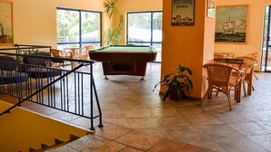 Una mesa de billar en una habitación con una escalera, sillas de mimbre y una planta en maceta.
