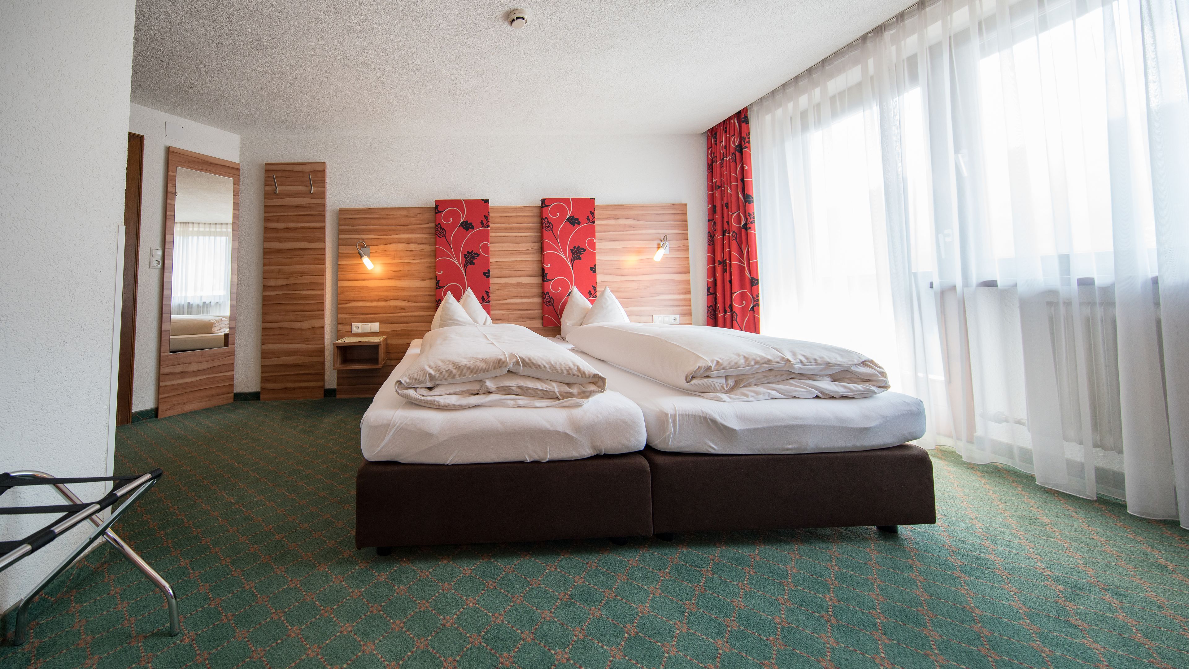 A hotel room with a large bed, a wooden headboard, a small bedside table, a mirror, and a window with sheer curtains.