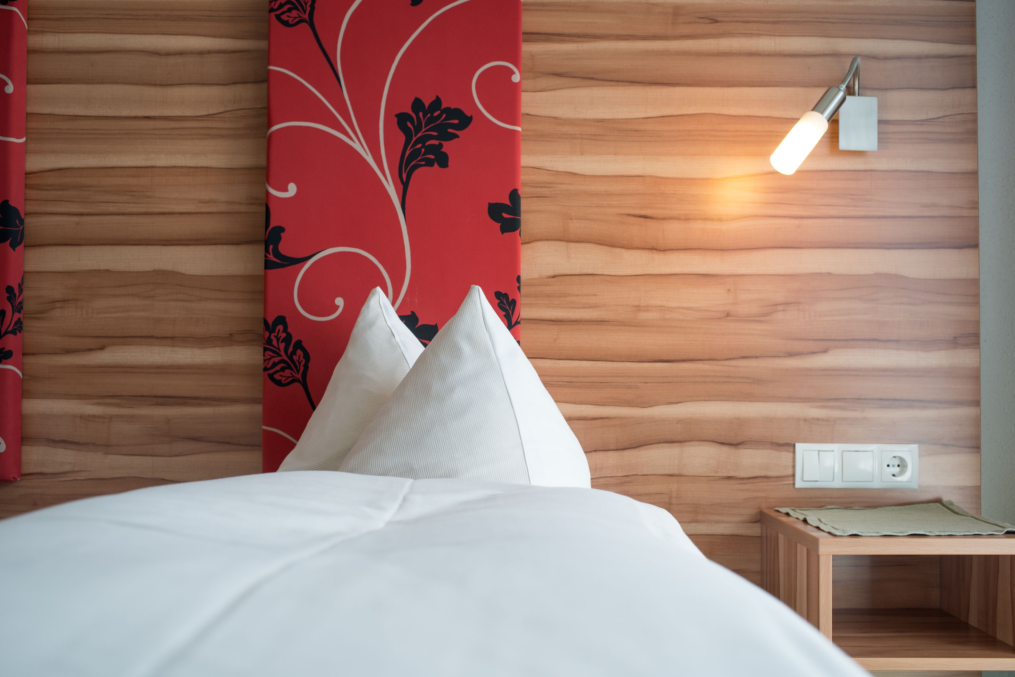 A neatly made bed with white bedding and decorative pillows, a wooden headboard with a red floral pattern, and a bedside table with a lamp.