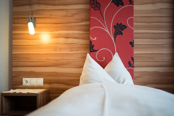 A neatly made bed with white bedding, a wooden headboard with a red floral pattern, a bedside table with a lamp, and a wall socket.