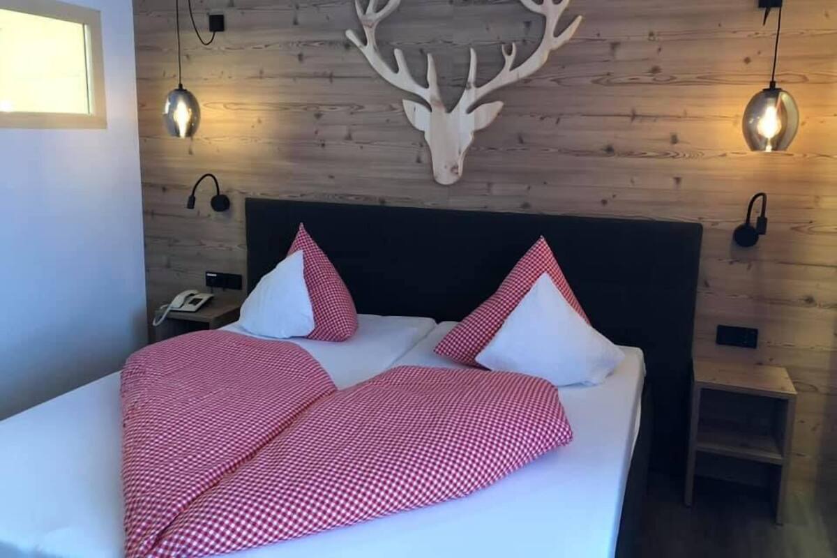 Een bed met een geruit deken, twee kussens en een houten muur met een opgeplakt hertenhoofd.