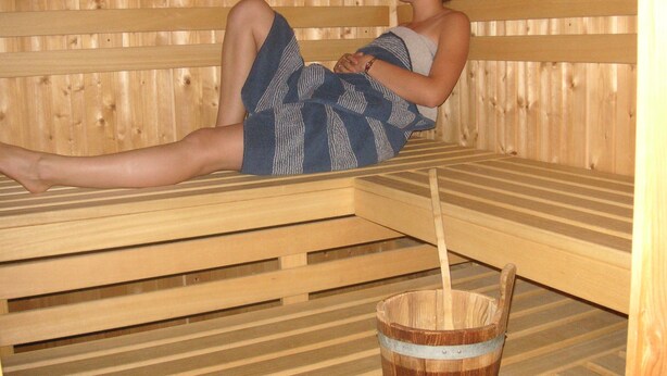 A person sitting on a wooden bench in a sauna room with a wooden bucket and a wooden bench in the foreground. - Gasthof Pension Posthansl (Heiterwang)