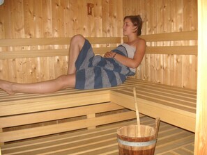 A person sitting on a wooden bench in a sauna room with a wooden bucket and a wooden bench in the foreground. - Gasthof Pension Posthansl (Heiterwang)
