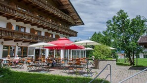 An outdoor seating area with tables and chairs, umbrellas, and a building with wooden balconies in the background. - Hotel Thaler (Thiersee)