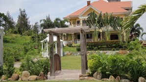 Une grande maison à deux étages, avec un toit recouvert de tuiles rouges, une véranda et un jardin bien entretenu agrémenté de rochers et de plantes tropicales.